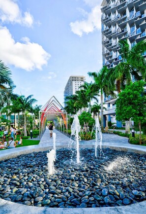 A modern building with a unique geometric facade, a central water feature with multiple fountains, and a pathway lined with palm trees. - Modern 1-bedroom apartment with AC in serene Doral (Doral)