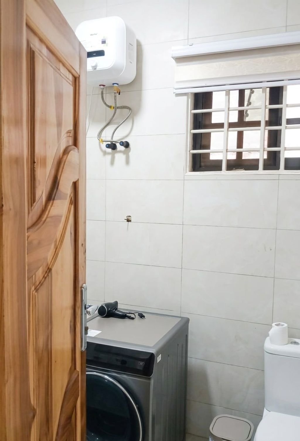 A compact bathroom with a washing machine, a toilet, and a window with bars.