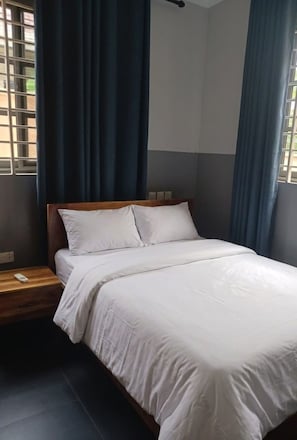 A bed with white bedding in a room with a dark floor, a wooden headboard, and a window with blue curtains. - 2-bedroom apartment in serene Greater Accra Region with AC (Greater Accra Region)