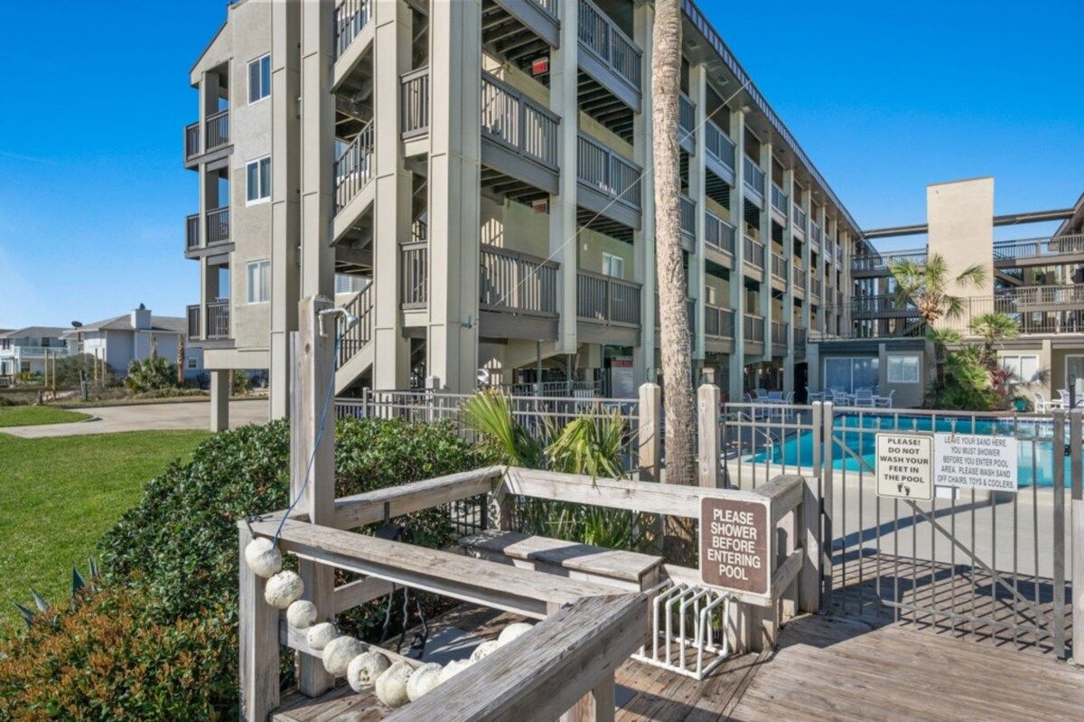 Un edificio de varios pisos con balcones, una zona de piscina y una terraza de madera con un letrero que indica ducharse antes de entrar a la piscina.
