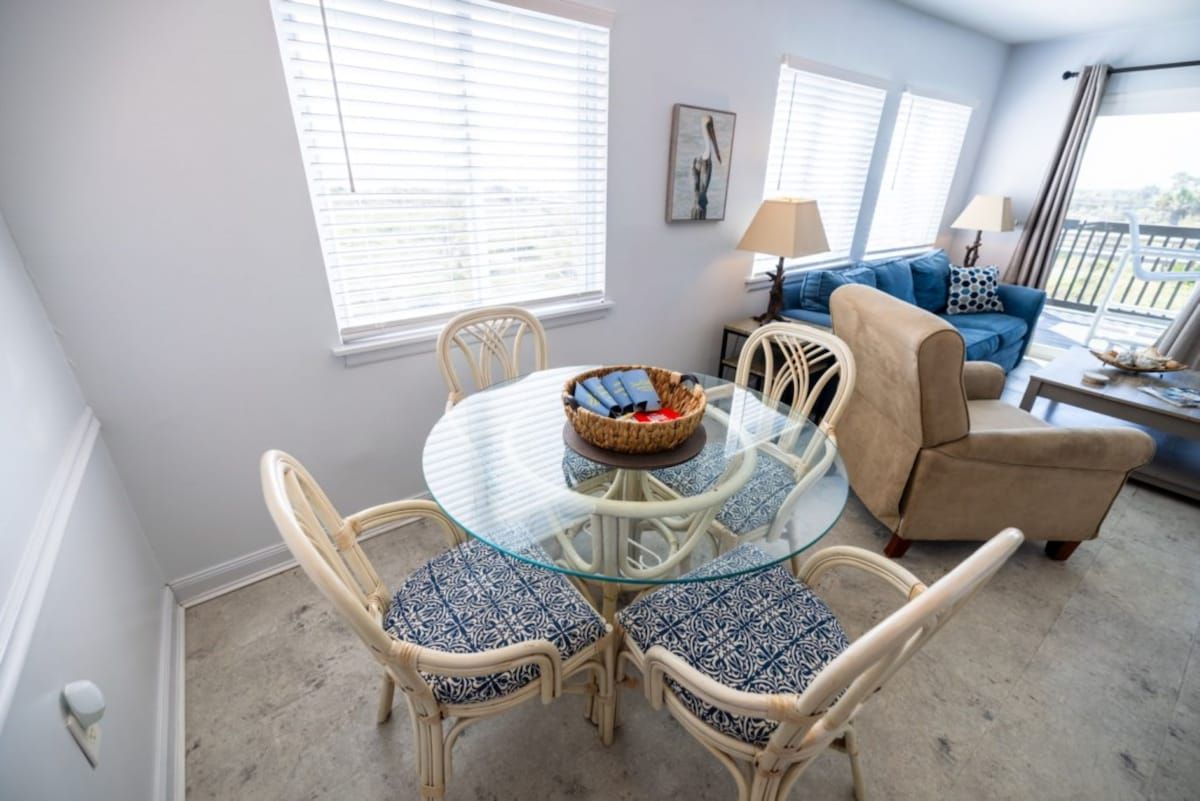 Une pièce comprenant une table ronde au plateau en verre, entourée de chaises en osier, d’un canapé beige et d’un fauteuil à motifs bleus.