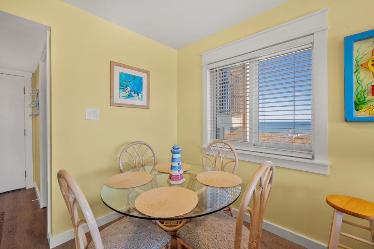 Un comedor con una mesa de vidrio, sillas de mimbre, un pequeño faro decorativo y una ventana con persianas.