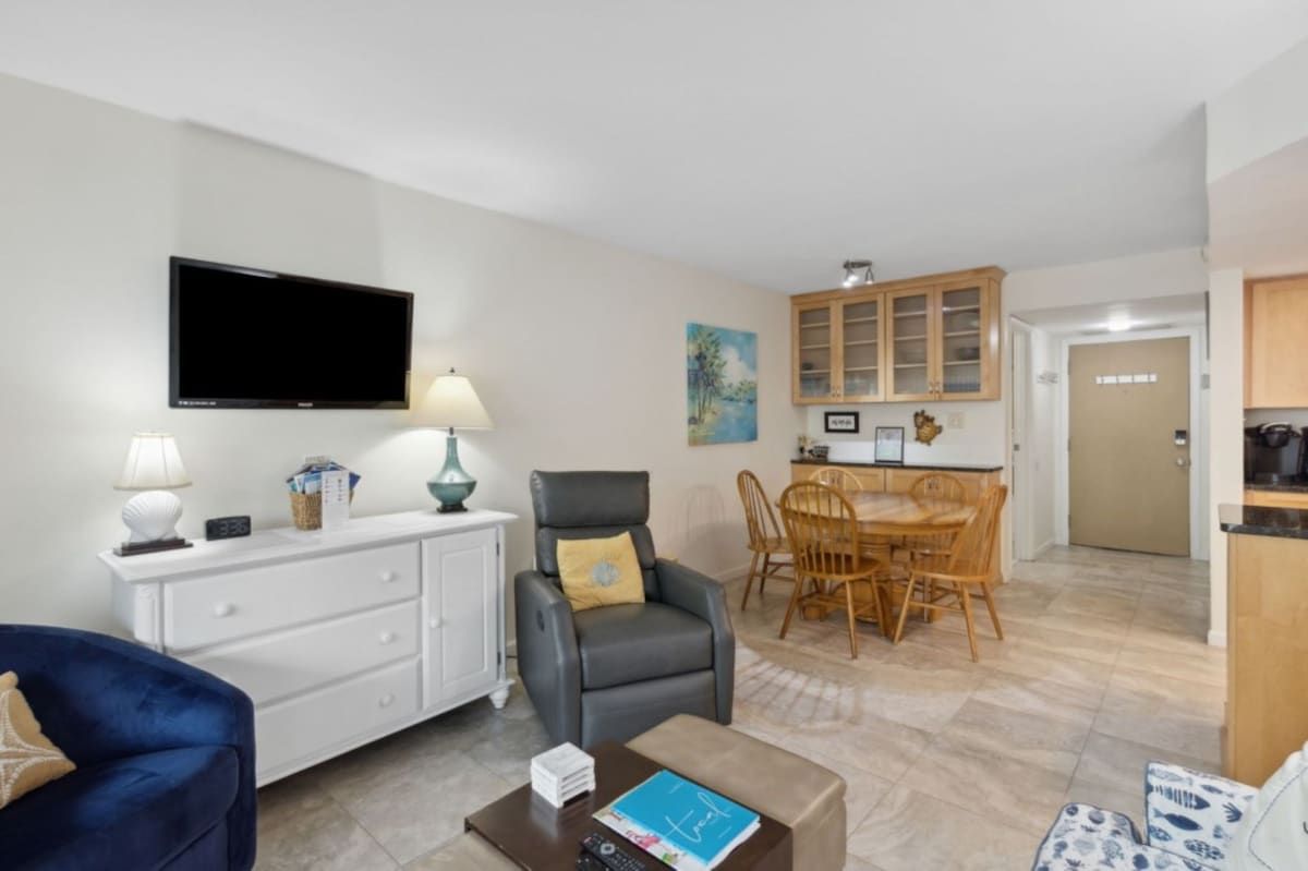 A living room with a flat-screen TV, a sofa, a coffee table, and a dining area with a wooden table and chairs.