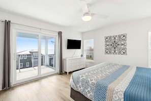 A bedroom with a large sliding glass door, a bed with patterned bedding, a white dresser, a flat-screen TV, and a decorative wall piece. - New 2025 • Ocean Views • Pool • 4BR (Fernandina Beach)