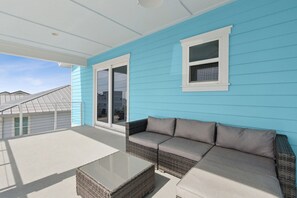 A covered outdoor seating area with a sofa and a glass-top table, a window, and a view of neighboring buildings. - Ocean View Beach House on Amelia Island with Private Elevator and Pool (Fernandina Beach)