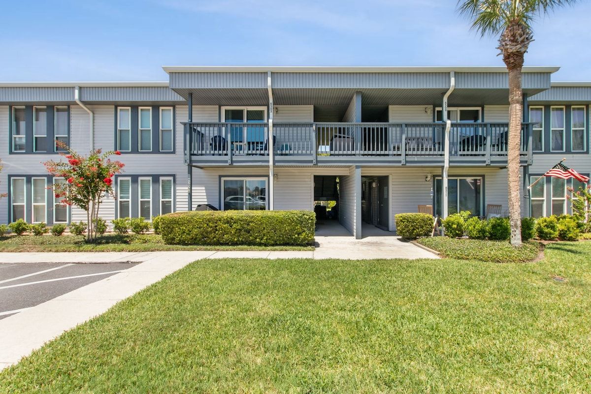 A multi-story residential building with balconies, a parking lot, and well-maintained lawns.