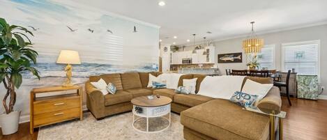 A living room with a sectional sofa, a coffee table, a ceiling fan, and a mural of the ocean.
