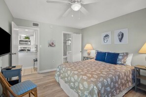 A bedroom with a bed, a desk, a chair, a ceiling fan, and a kitchenette in the background. - Top Floor • Beach Access • Pool • Amelia (Fernandina Beach)