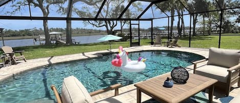 A covered outdoor swimming pool area with lounge chairs and a table, surrounded by trees and a view of a lake.