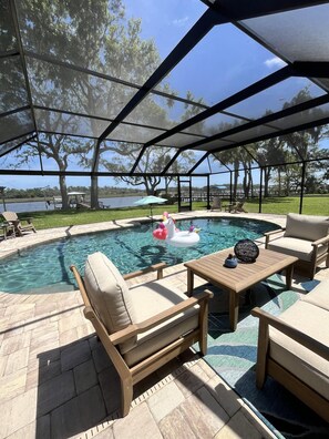 A covered outdoor swimming pool area with lounge chairs and a table, surrounded by trees and a view of a lake. - Spacious 3BR home with private pool and water views in Yulee. Easy access to Amelia Island. (Yulee)