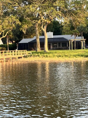 A lakeside property with a covered outdoor seating area, a building with large windows, and a wooden deck. - Spacious 3BR home with private pool and water views in Yulee. Easy access to Amelia Island. (Yulee)