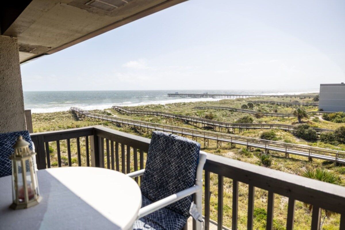 Un balcon avec une table et une chaise, donnant sur un paysage côtier avec une jetée et un sentier pédestre.