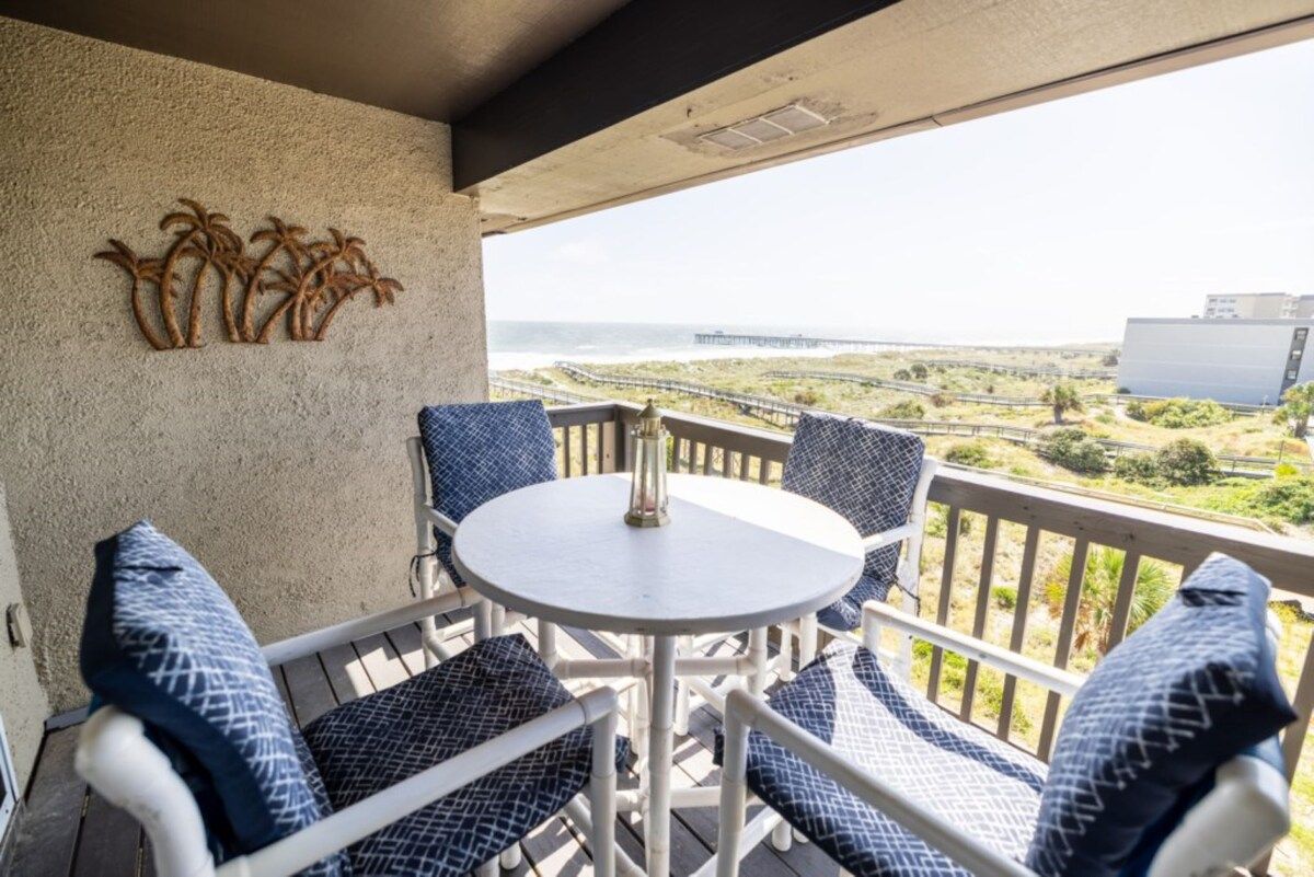 A balcony with a white table and blue patterned chairs, overlooking a coastal view with a pier and buildings.