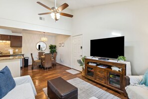 Ein modernes Wohnzimmer mit Flachbildfernseher, Deckenventilator, Essbereich mit rundem Tisch und Stühlen sowie Küche mit Backsteinwand.
