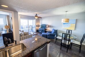 A modern kitchen with a granite countertop, stainless steel sink, and a built-in dishwasher. There is a dining area with a round table and chairs, and a living room with a sofa and a television. - Oceanfront Amelia Island Condo with Ocean View Balcony, Elevator, and Pool (Fernandina Beach)