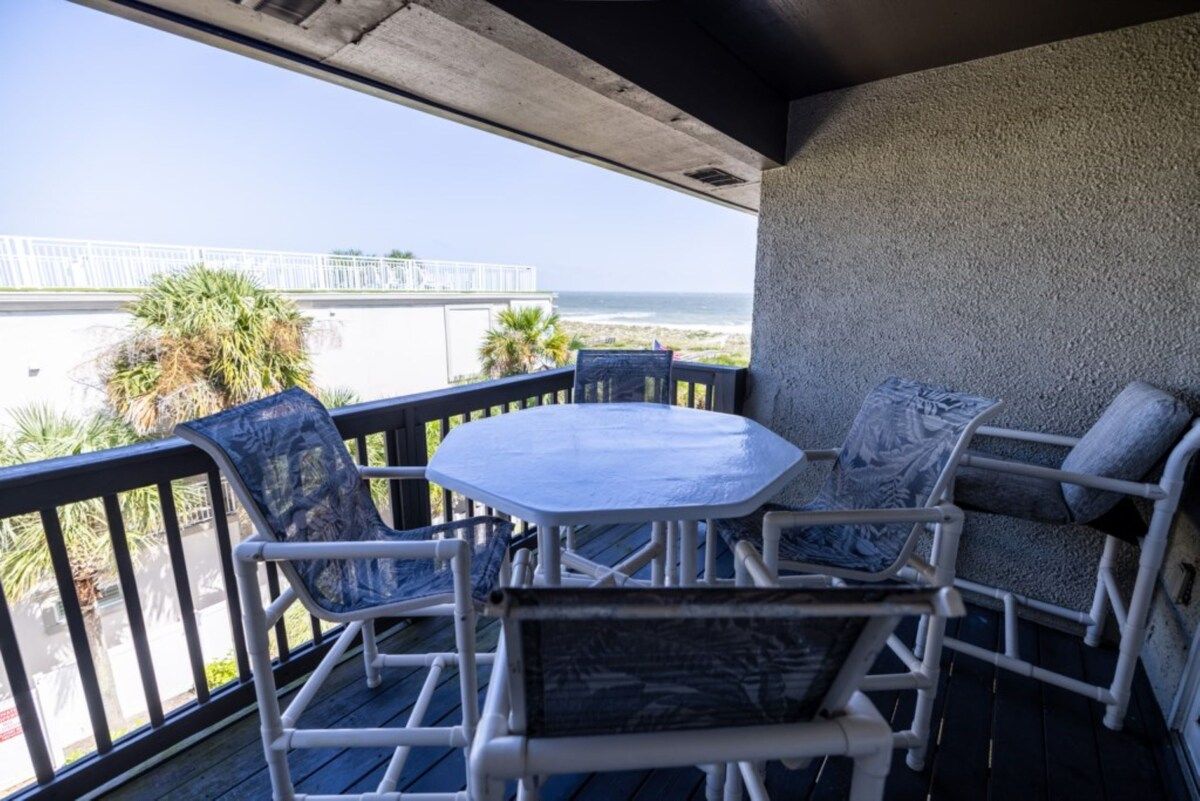 Un balcon avec une table et des chaises, donnant sur une plage et une clôture blanche.