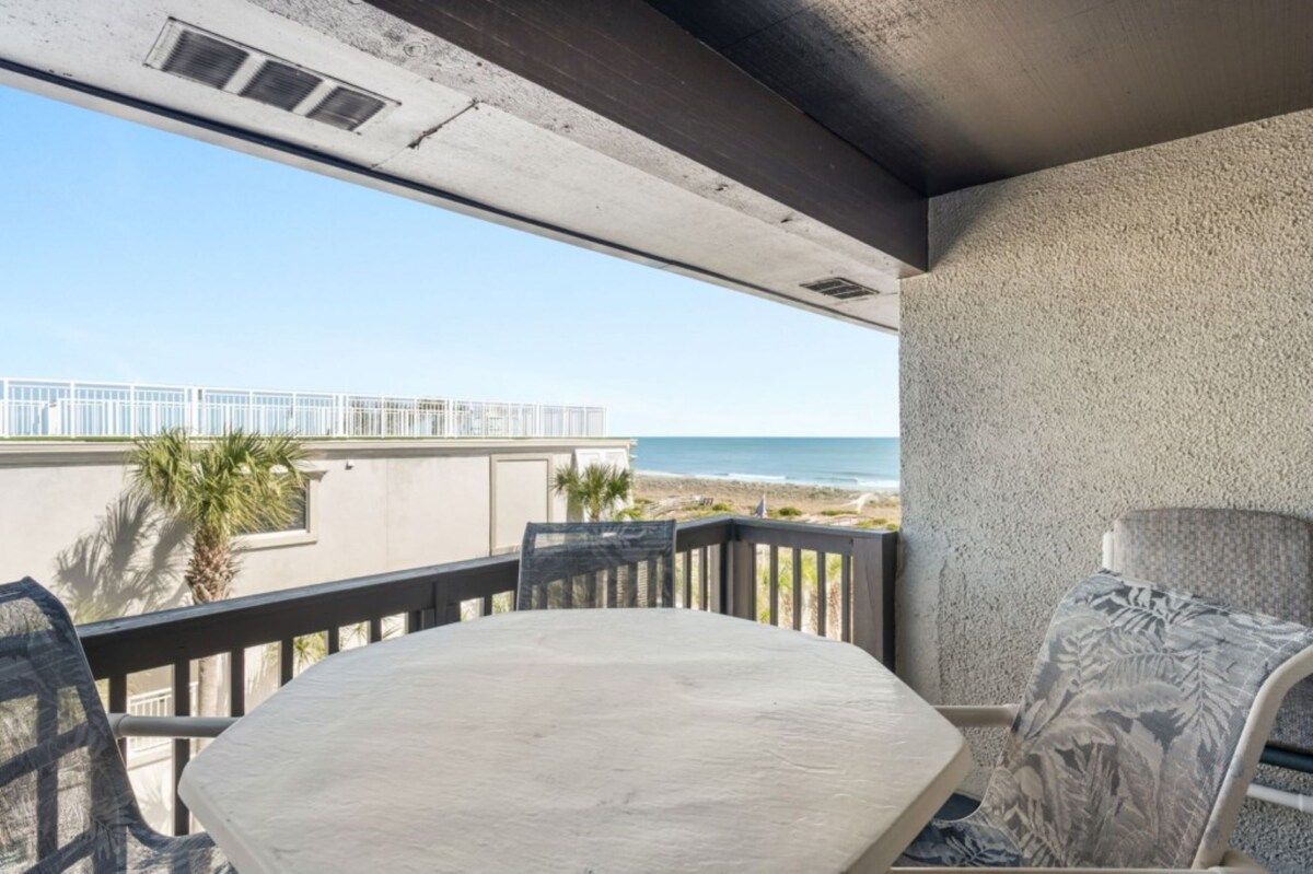 Een balkon met een tafel en stoelen, met uitzicht op een strand en een gebouw.