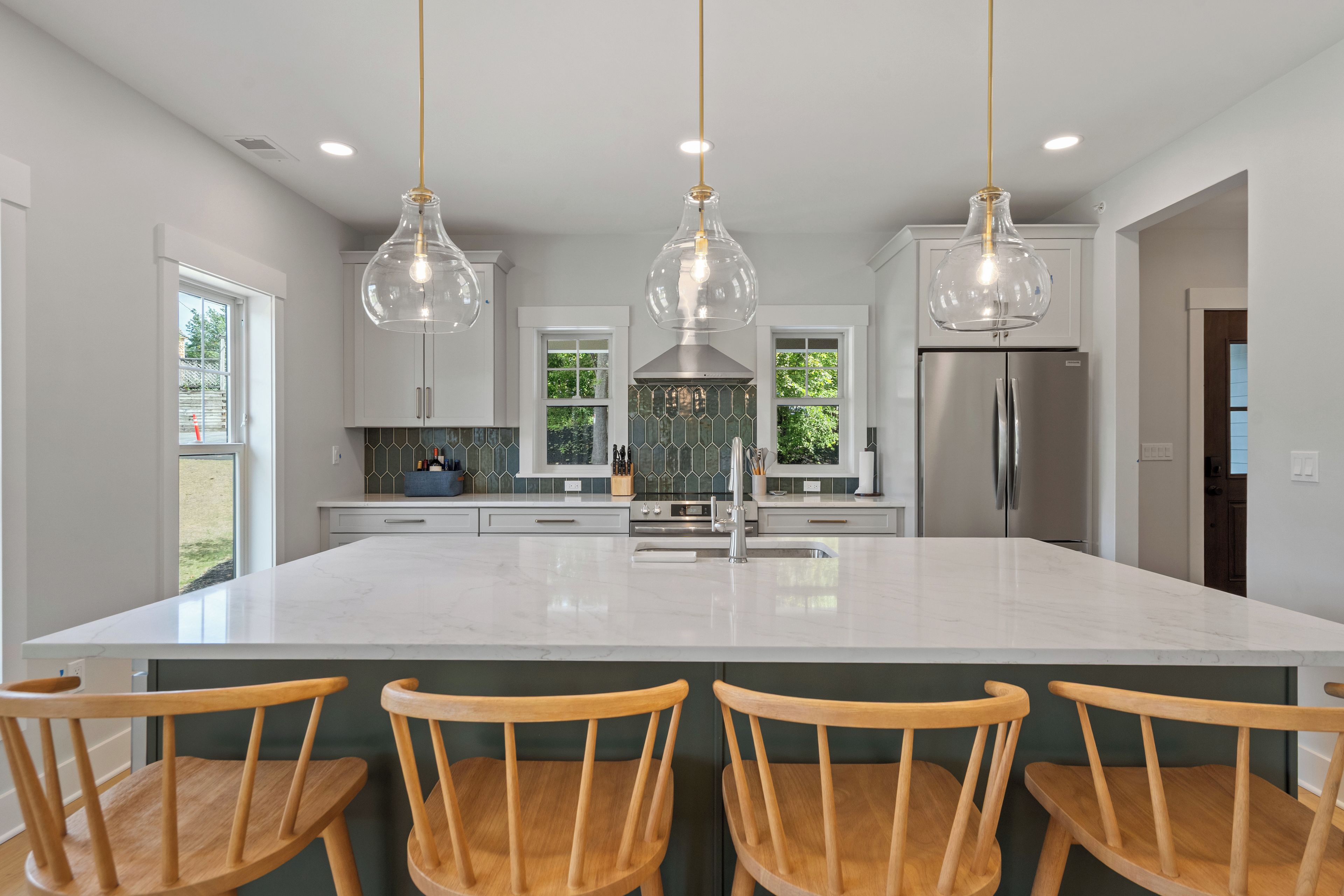 A modern kitchen with a large island, pendant lights, and wooden chairs.