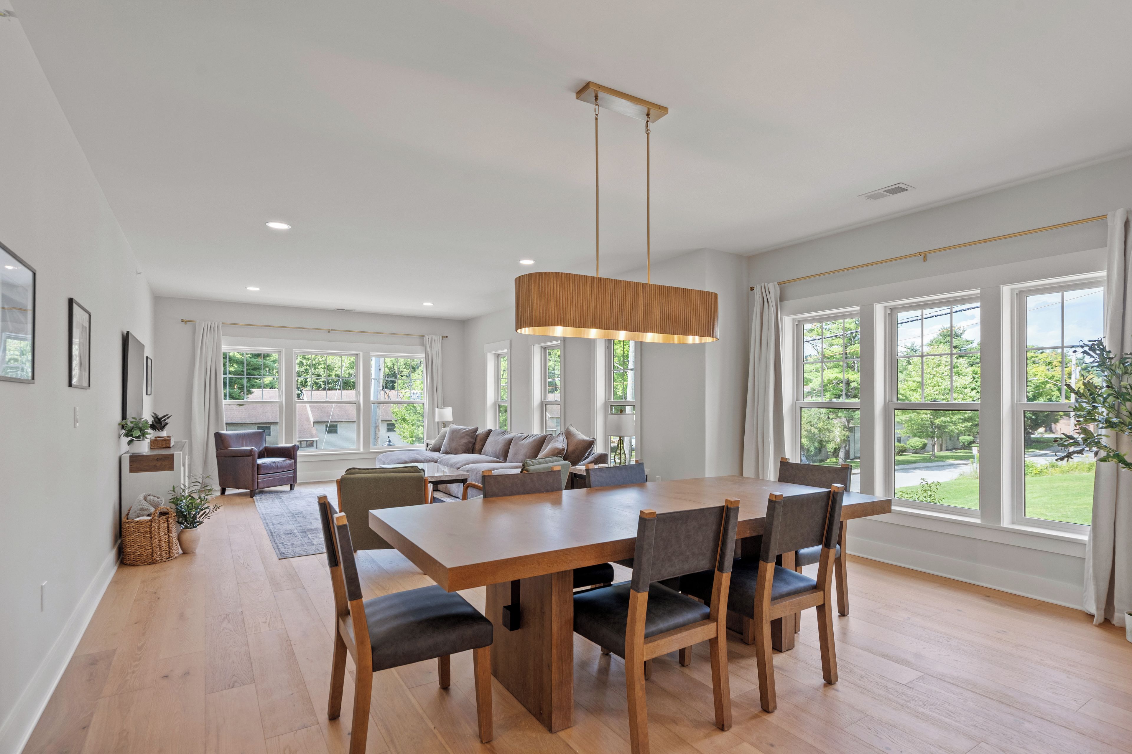 A spacious dining area with a wooden table and chairs, large windows with a view of greenery, and a modern pendant light.