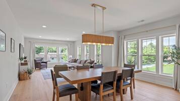 A spacious dining area with a wooden table and chairs, large windows with a view of greenery, and a modern pendant light.