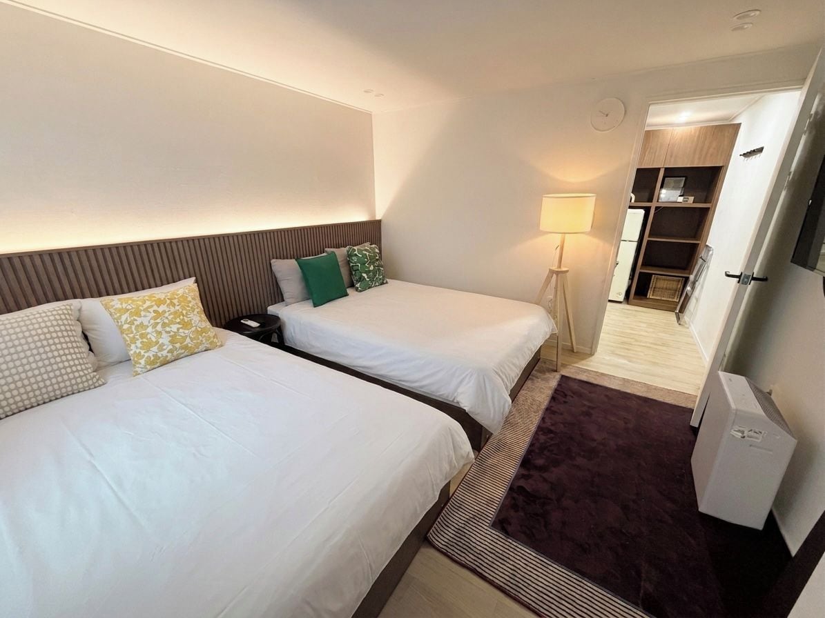 A hotel room with two beds, a bedside table, a lamp, a wardrobe, and a refrigerator.