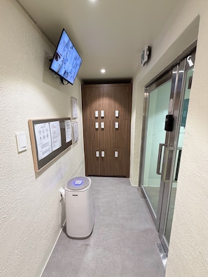 Corredor com televisão na parede, um mural de avisos, um alto-falante branco e uma porta com painel de vidro.