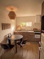 Una cocina compacta con una mesa redonda para comer, dos sillas, una lámpara colgante, una ventana con plantas y un microondas.