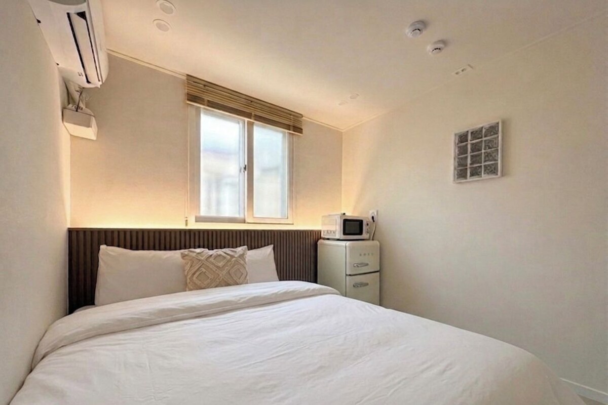 Quarto de hotel com cama, ar-condicionado, janela com persianas, micro-ondas e aquecedor de parede.