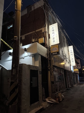 Um edifício com várias placas, incluindo uma com texto em coreano e outra com um número de telefone. Fios elétricos e uma parede de tijolos são visíveis.