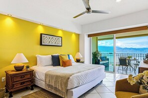 A bedroom with a bed, bedside lamps, a ceiling fan, a framed picture, and a balcony with a view. - Beachfront-2 BR Plus 2 BR-Apartment Shared Balcony (Puerto Vallarta)