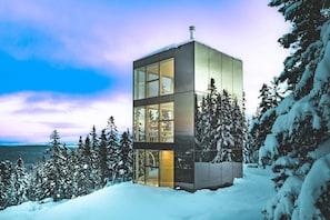 A modern, cube-shaped building with large glass windows, surrounded by snow-covered trees and a body of water in the background. - BLACK MIRROR / Glass Retreat Above the Mountains (Capitale-Nationale)