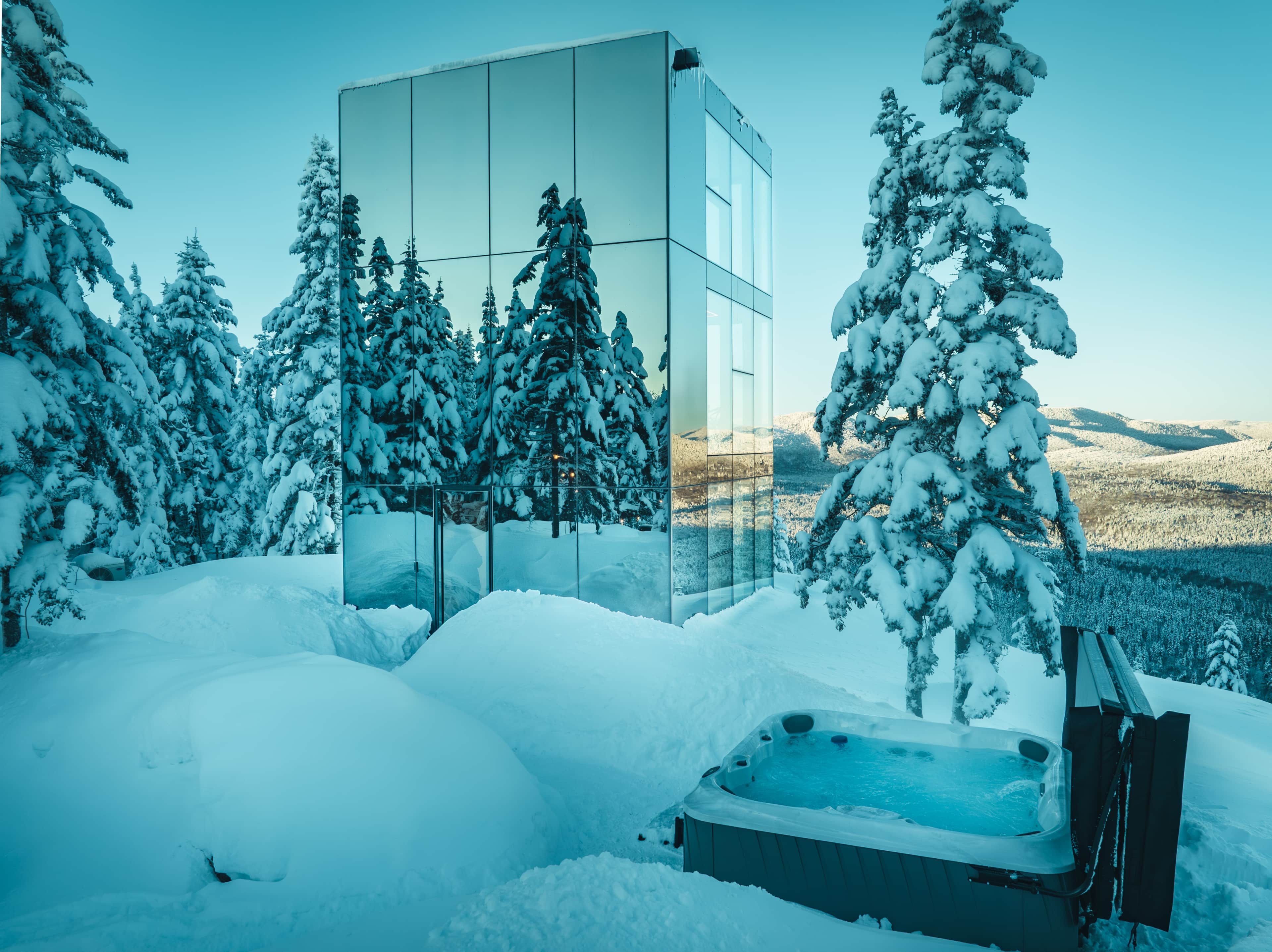 Um edifício moderno de vidro rodeado por árvores cobertas de neve e uma banheira de hidromassagem em uma paisagem nevada.