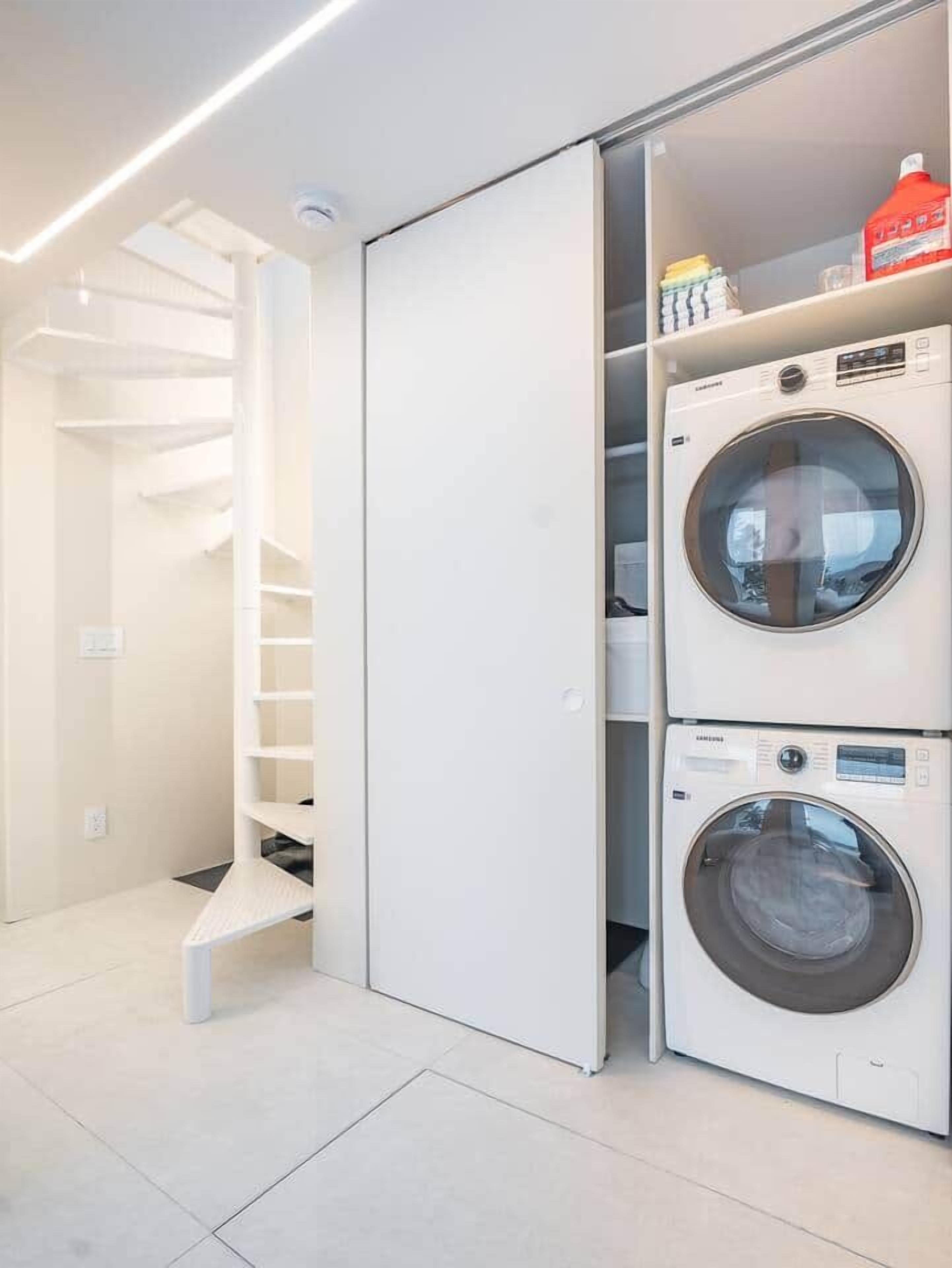 A laundry room with two stacked washing machines and a white shelving unit with folded clothes.