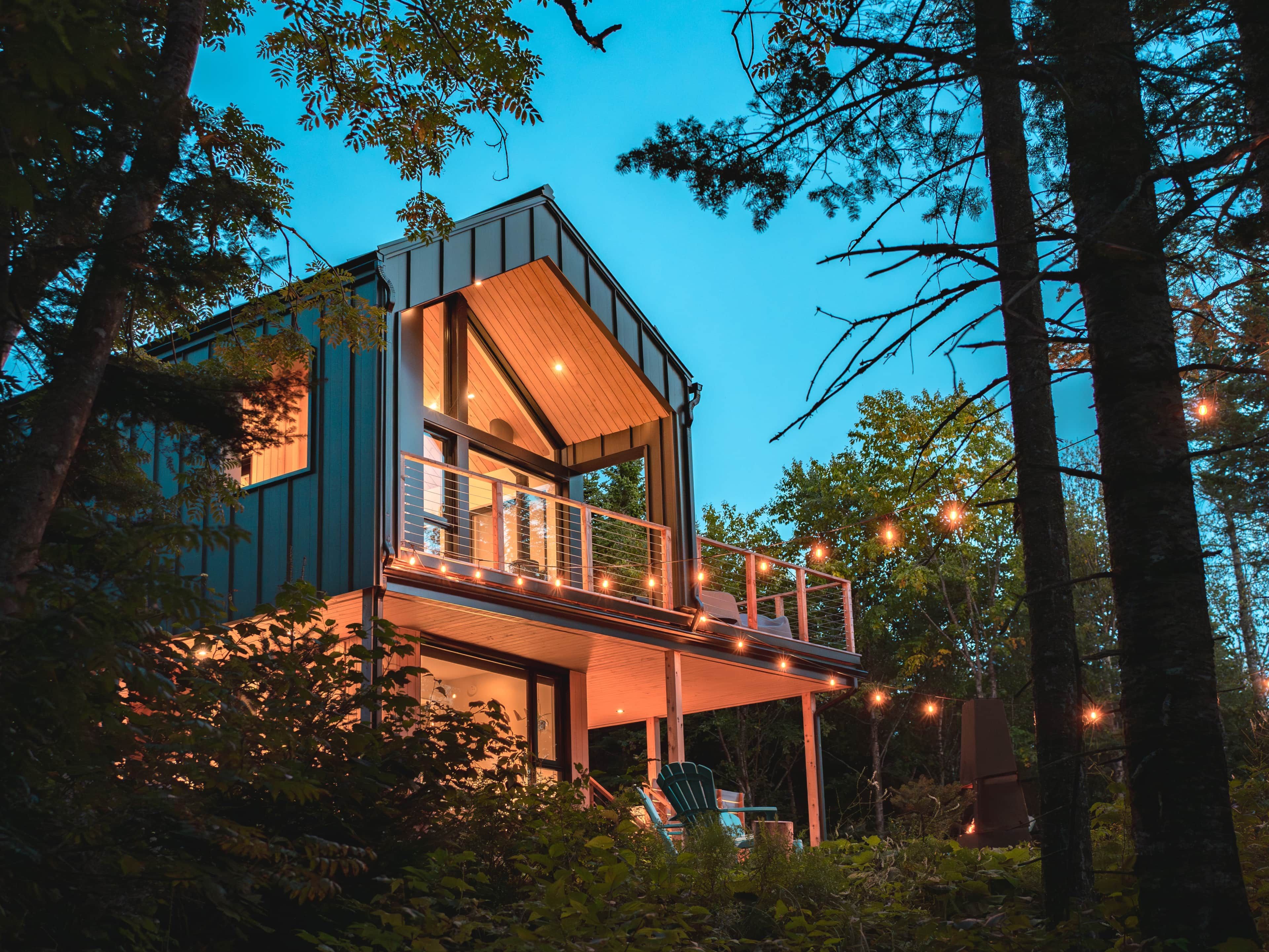 Une cabane moderne à deux étages, dotée d’un balcon et entourée d’arbres.