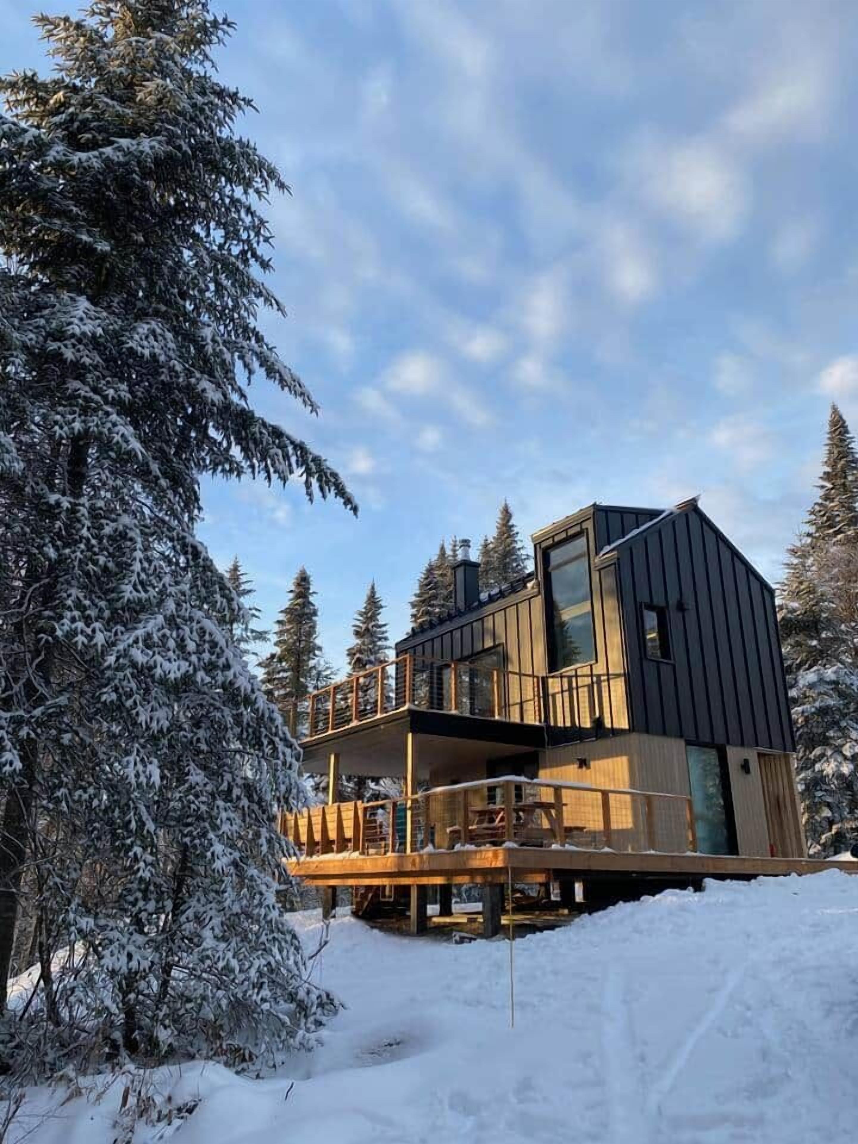 Moderni mökki, jossa on puinen terassi ja lasinen parveke, ja sen ympärillä on lumipeitteisiä puita.