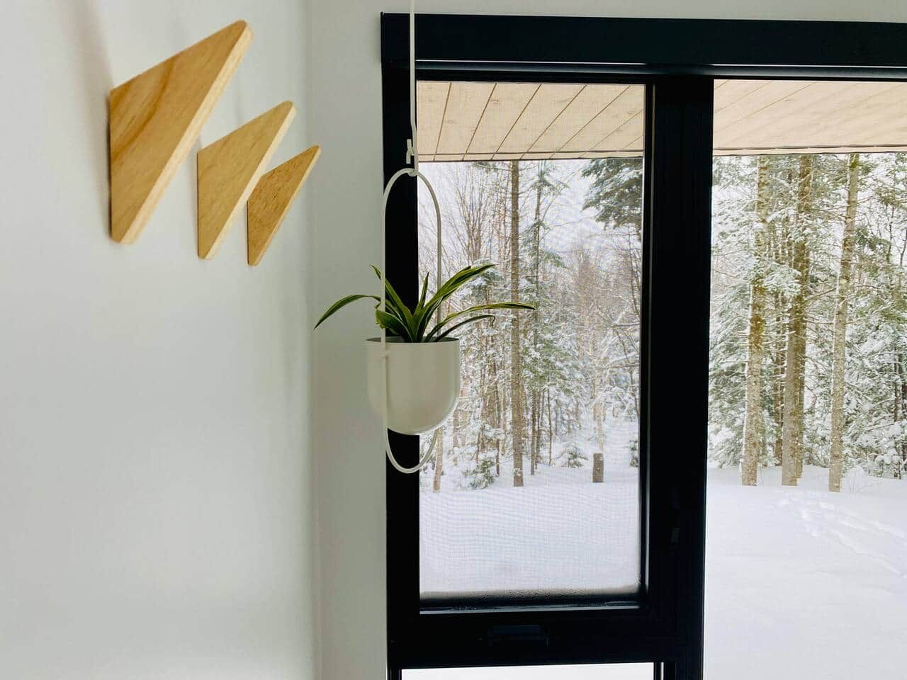 Ett fönster med utsikt över en snötäckt skog, en hängande växt och geometriska väggdekorationer.