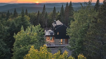 En stuga med en veranda och en eldstad, omgiven av tät skog och med berg i bakgrunden.