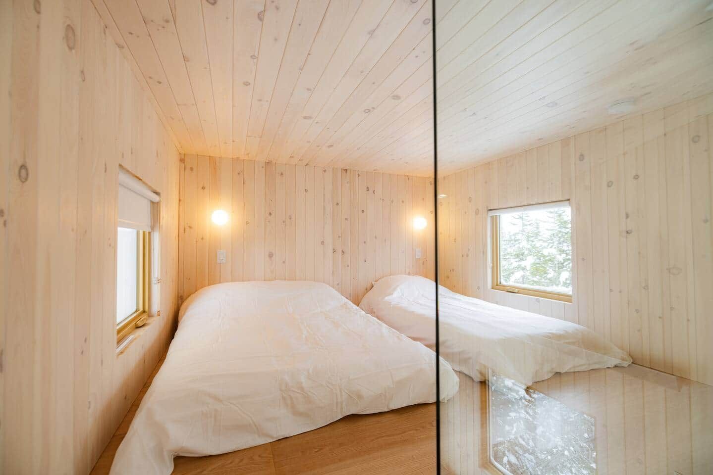 Una habitación con paneles de madera, dos camas, un ventanal y una lámpara fijada a la pared.