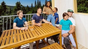 Un groupe de personnes est assis autour d’une table en bois sur un balcon d’où l’on peut admirer des arbres et un ciel dégagé.