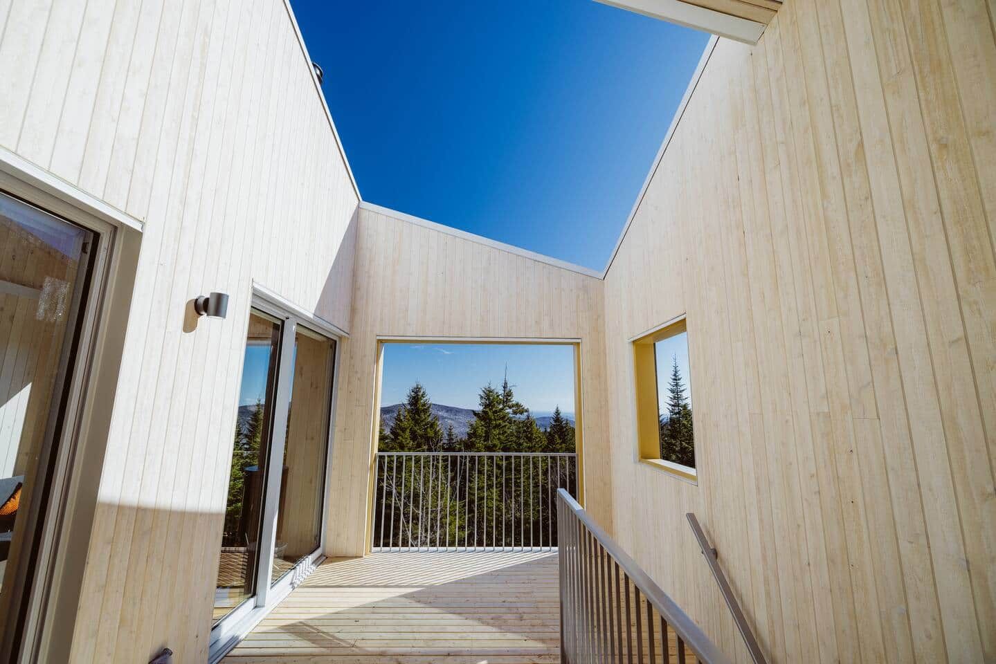 A modern building with a wooden exterior, a balcony with a metal railing, and a clear view of a forested mountain range.