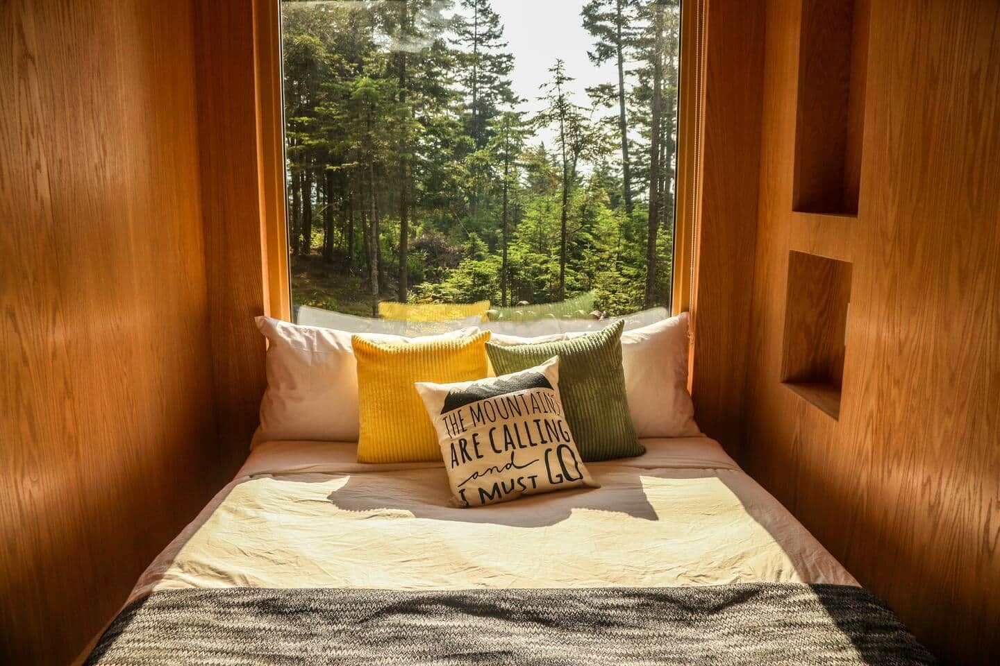 Acogedor cuarto tipo cabaña con asiento junto a la ventana, almohadones y vista a un bosque.