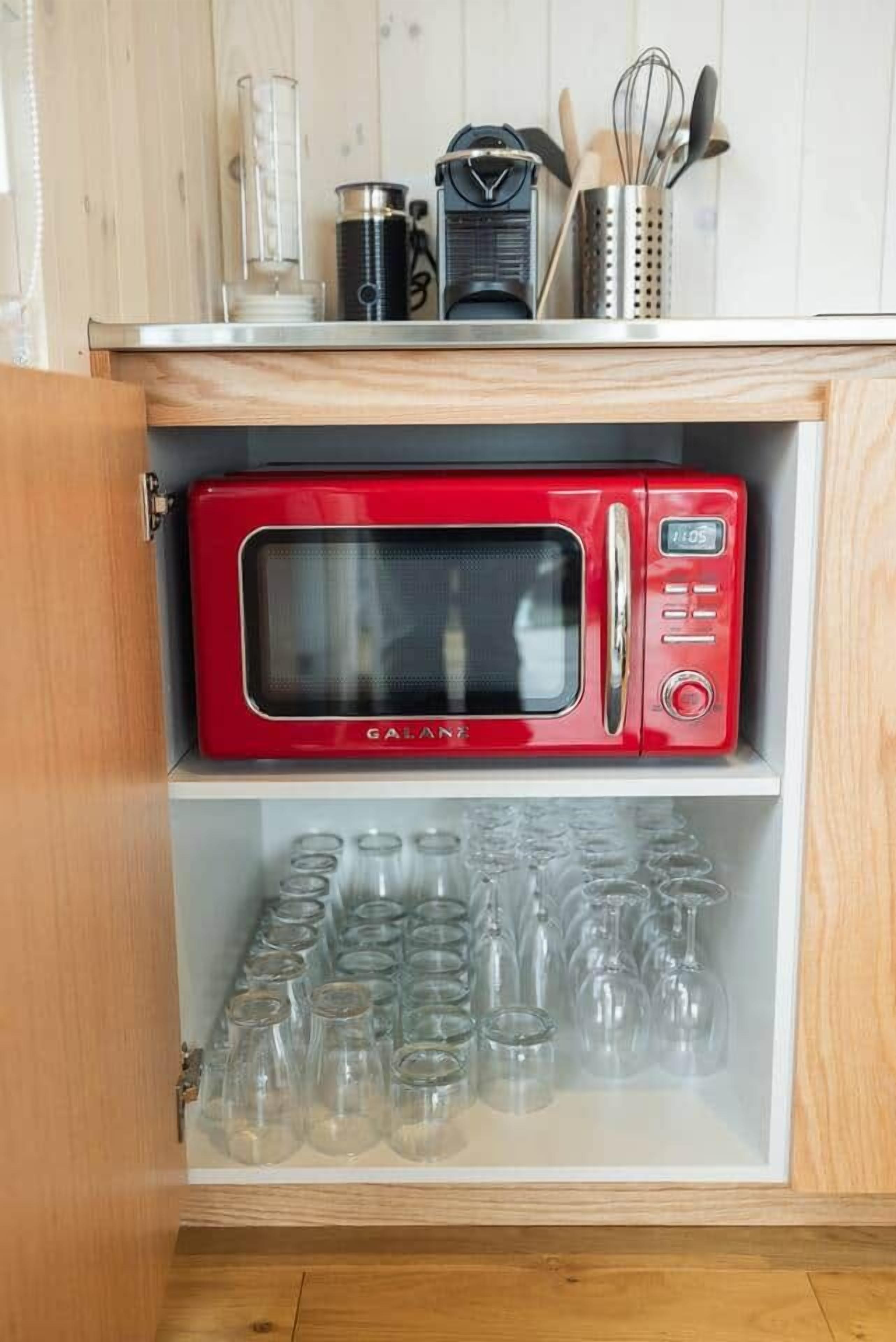 A kitchen with a red microwave on a wooden cabinet shelf, a glass container, and a metal container with utensils.
