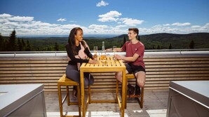 To personer sidder ved et træbord på en udendørs terrasse med en smuk udsigt over bjerge og træer.