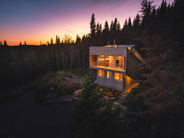A modern two-story cabin with a balcony, surrounded by a forested landscape.
