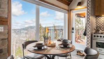 Espaço de refeições com mesa posta para dois, vista para o exterior e cozinha equipada com fogão.