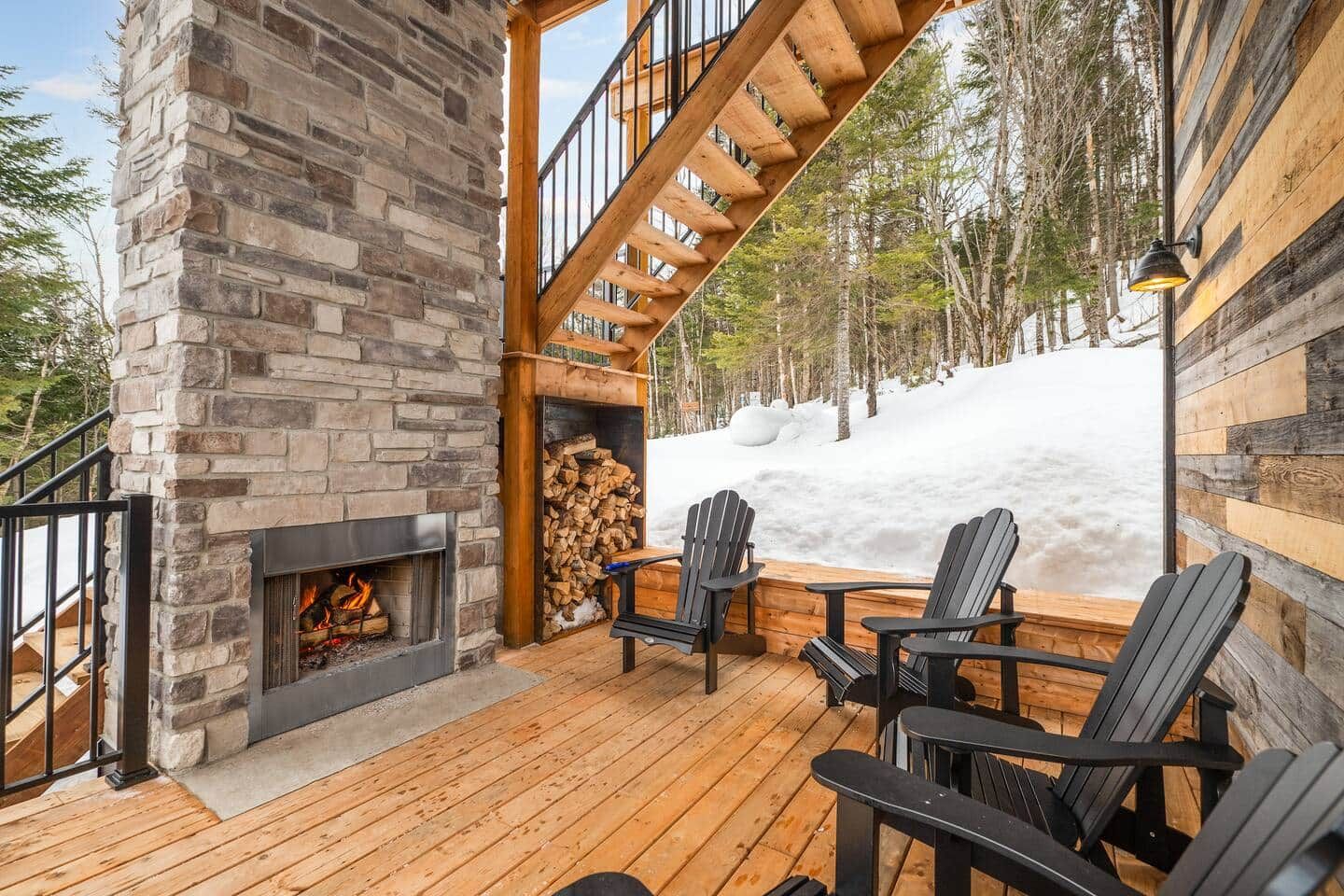 Une cheminée en pierre avec un feu de bois, une terrasse en bois, des fauteuils Adirondack et une vue sur un paysage enneigé.