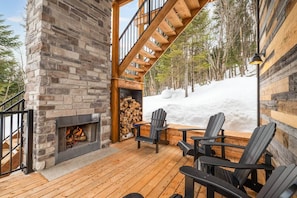 A stone fireplace with a fire, wooden deck, Adirondack chairs, and a view of a snowy landscape. - CHAMONIX / Micro-Chalet / Mountain Escape near Quebec City (Capitale-Nationale)
