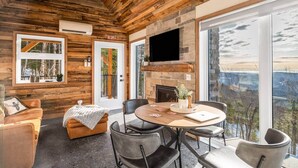 A log cabin interior with a stone fireplace, a television mounted on the wall, and a dining area with a view of the outdoors. - CHAMONIX / Micro-Chalet / Mountain Escape near Quebec City (Capitale-Nationale)