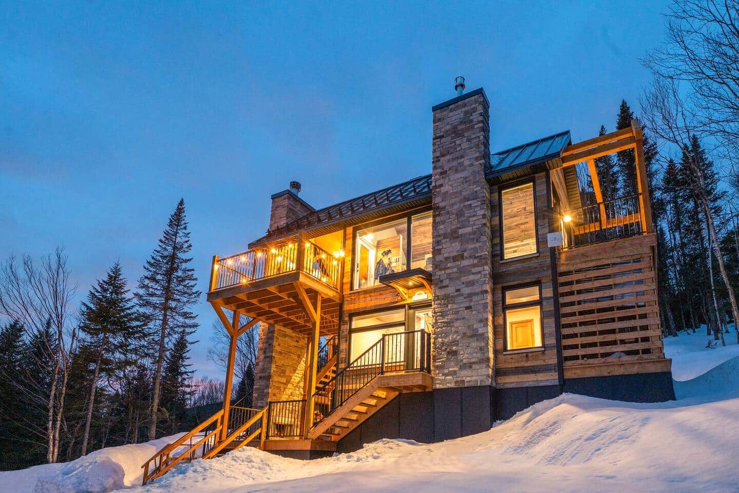 Una cabaña de madera de varios niveles con un balcón, rodeada de nieve y árboles.