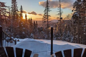 A snow-covered deck with a railing, surrounded by tall evergreen trees and a sunset in the background.